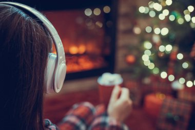 Pijamalı bir kadın, elinde bir fincan sıcak kakao ve marshmallow ile kış akşamı şömine ateşi ve noel ağacının yanında oturup ısınıyor..