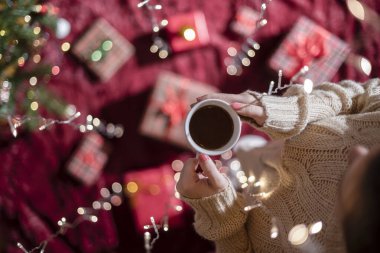 Elinde bir fincan sıcak çikolata ve aile için Noel hediyelerini Noel ağacının yanında yumuşak ekose kumaşla paketleyen bir kadın. Parlayan çelenk boyunca en üst manzara.