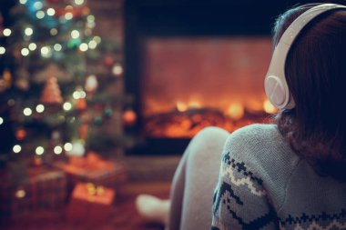Kulaklıklı kadın kış akşamı şömine ateşi ve yılbaşı ağacının yanında oturup ısınıyor..