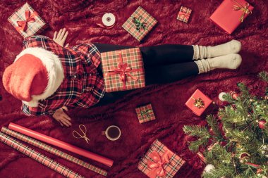 Dizlerinin üzerinde Noel hediyesi kutusu olan bir kadının en iyi görüntüsü. Noel ağacının yanındaki kabarık ekose üzerinde oturan aile için kadın paketleme hediyeleri. Düz yatıyordu.