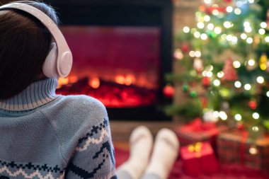 Yanlız üzgün kulaklıklı kadın kış akşamı şömine ateşi ve yılbaşı ağacının yanında oturup ısınıyor..