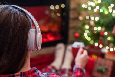 Pijamalı bir kadın, elinde bir fincan sıcak kakao ve marshmallow ile kış akşamı şömine ateşi ve noel ağacının yanında oturup ısınıyor..