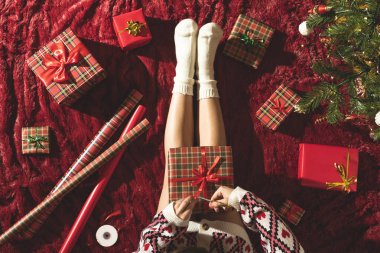 Dizlerinin üzerinde Noel hediyesi kutusu olan bir kadının en iyi görüntüsü. Noel ağacının yanındaki kabarık ekose üzerinde oturan aile için kadın paketleme hediyeleri. Düz yatıyordu.