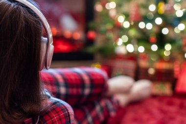 Kulaklıklı, pijamalı bir kadın kış akşamı şömine ateşi ve noel ağacının yanında yatıp ısınıyor..