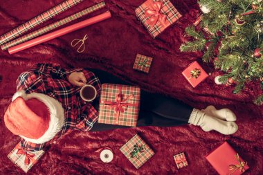 Dizlerinin üzerinde Noel hediyesi kutusu olan bir kadının en iyi görüntüsü. Noel ağacının yanındaki kabarık ekose üzerinde oturan aile için kadın paketleme hediyeleri. Düz yatıyordu.