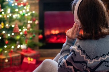 Yanlız üzgün kulaklıklı kadın kış akşamı şömine ateşi ve yılbaşı ağacının yanında oturup ısınıyor..