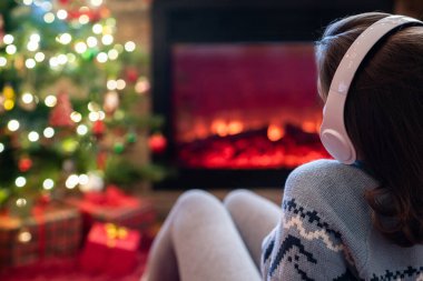 Kulaklıklı kadın kış akşamı şömine ateşi ve yılbaşı ağacının yanında oturup ısınıyor..