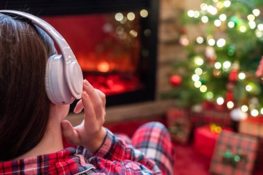 Kulaklıklı bir kadın kış akşamı şömine ateşi ve noel ağacının yanında oturup ısınıyor..