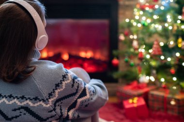 Yanlız üzgün kulaklıklı kadın kış akşamı şömine ateşi ve yılbaşı ağacının yanında oturup ısınıyor..