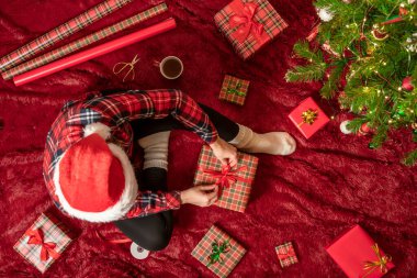 Kırmızı Noel Baba şapkalı bir kadının, Noel ağacının yanındaki kabarık ekose kumaşta oturan aile için Noel hediyesi kutularını paketlediği en iyi manzara. Düz yatıyordu.