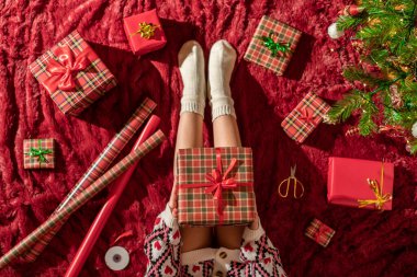 Dizlerinin üzerinde Noel hediyesi kutusu olan bir kadının en iyi görüntüsü. Noel ağacının yanındaki kabarık ekose üzerinde oturan aile için kadın paketleme hediyeleri. Düz yatıyordu.