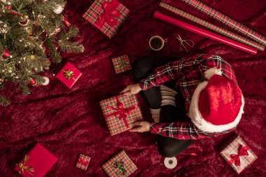 Kırmızı Noel Baba şapkalı bir kadının, Noel ağacının yanındaki kabarık ekose kumaşta oturan aile için Noel hediyesi kutularını paketlediği en iyi manzara. Düz yatıyordu.