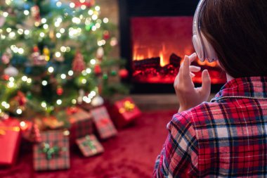 Yanlız üzgün kulaklıklı kadın kış akşamı şömine ateşi ve yılbaşı ağacının yanında oturup ısınıyor..