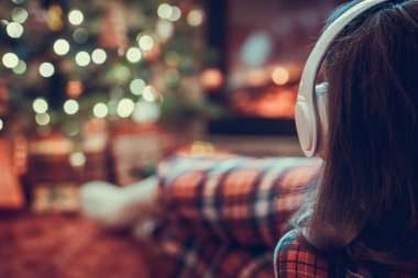 Kulaklıklı, pijamalı bir kadın kış akşamı şömine ateşi ve noel ağacının yanında yatıp ısınıyor..