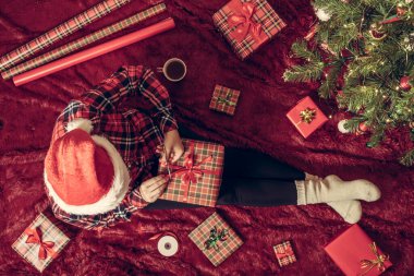 Dizlerinin üzerinde Noel hediyesi kutusu olan bir kadının en iyi görüntüsü. Noel ağacının yanındaki kabarık ekose üzerinde oturan aile için kadın paketleme hediyeleri. Düz yatıyordu.