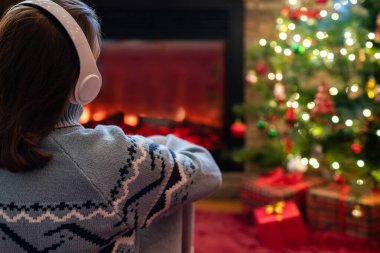 Kulaklıklı kadın kış akşamı şömine ateşi ve yılbaşı ağacının yanında oturup ısınıyor..
