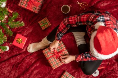 Kırmızı Noel Baba şapkalı bir kadının, Noel ağacının yanındaki kabarık ekose kumaşta oturan aile için Noel hediyesi kutularını paketlediği en iyi manzara. Düz yatıyordu.