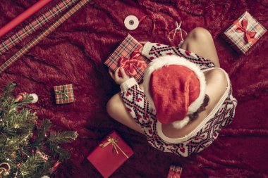 Kırmızı Noel Baba şapkalı bir kadının, Noel ağacının yanındaki kabarık ekose kumaşta oturan aile için Noel hediyesi kutularını paketlediği en iyi manzara. Düz yatıyordu.