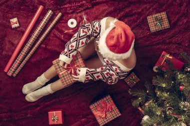 Dizlerinin üzerinde Noel hediyesi kutusu olan bir kadının en iyi görüntüsü. Noel ağacının yanındaki kabarık ekose üzerinde oturan aile için kadın paketleme hediyeleri. Düz yatıyordu.