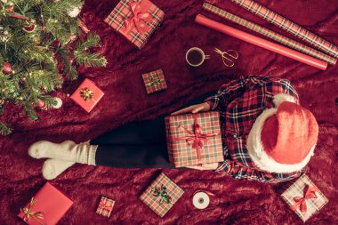 Dizlerinin üzerinde Noel hediyesi kutusu olan bir kadının en iyi görüntüsü. Noel ağacının yanındaki kabarık ekose üzerinde oturan aile için kadın paketleme hediyeleri. Düz yatıyordu.