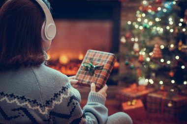 Elinde Noel hediyesi kutusu olan kulaklıklı bir kadın şömine ve noel ağacının yanında, yumuşak ekose üzerinde oturuyor.. 