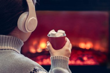 Kulaklıklı, elinde sıcak kakao ve lokum olan bir kadın kış akşamı şömine ateşi ve noel ağacının yanında oturup ısınıyor..