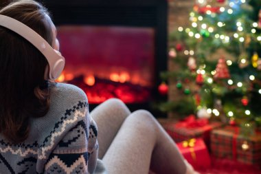 Kulaklıklı kadın kış akşamı şömine ateşi ve yılbaşı ağacının yanında oturup ısınıyor..