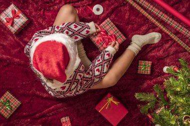 Kırmızı Noel Baba şapkalı bir kadının, Noel ağacının yanındaki kabarık ekose kumaşta oturan aile için Noel hediyesi kutularını paketlediği en iyi manzara. Düz yatıyordu.