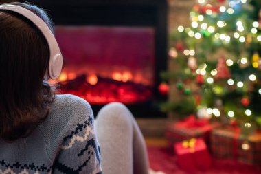 Yanlız üzgün kulaklıklı kadın kış akşamı şömine ateşi ve yılbaşı ağacının yanında oturup ısınıyor..