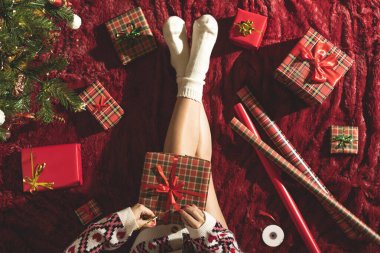 Dizlerinin üzerinde Noel hediyesi kutusu olan bir kadının en iyi görüntüsü. Noel ağacının yanındaki kabarık ekose üzerinde oturan aile için kadın paketleme hediyeleri. Düz yatıyordu.