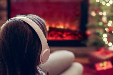 Kulaklıklı kadın kış akşamı şömine ateşi ve yılbaşı ağacının yanında oturup ısınıyor..