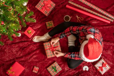 Kırmızı Noel Baba şapkalı bir kadının, Noel ağacının yanındaki kabarık ekose kumaşta oturan aile için Noel hediyesi kutularını paketlediği en iyi manzara. Düz yatıyordu.