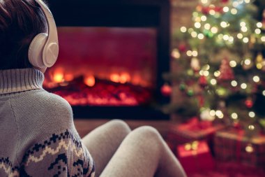 Kulaklıklı kadın kış akşamı şömine ateşi ve yılbaşı ağacının yanında oturup ısınıyor..
