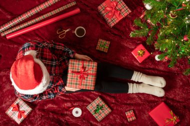 Dizlerinin üzerinde Noel hediyesi kutusu olan bir kadının en iyi görüntüsü. Noel ağacının yanındaki kabarık ekose üzerinde oturan aile için kadın paketleme hediyeleri. Düz yatıyordu.