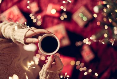 Elinde bir fincan sıcak çikolata ve aile için Noel hediyelerini Noel ağacının yanında yumuşak ekose kumaşla paketleyen bir kadın. Parlayan çelenk boyunca en üst manzara.
