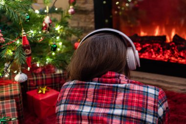 Kulaklıklı bir kadın kış akşamı şömine ateşi ve noel ağacının yanında oturup ısınıyor..