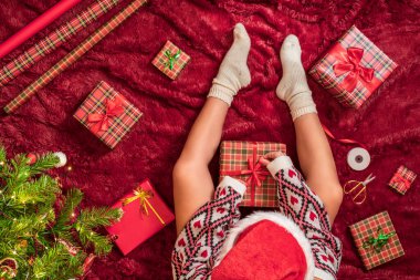 Kırmızı Noel Baba şapkalı bir kadının, Noel ağacının yanındaki kabarık ekose kumaşta oturan aile için Noel hediyesi kutularını paketlediği en iyi manzara. Düz yatıyordu.