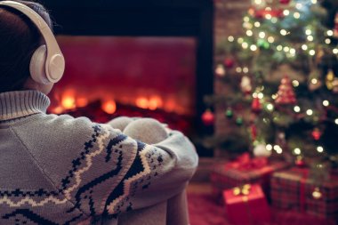 Yanlız üzgün kulaklıklı kadın kış akşamı şömine ateşi ve yılbaşı ağacının yanında oturup ısınıyor..