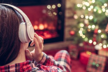 Kulaklıklı bir kadın kış akşamı şömine ateşi ve noel ağacının yanında oturup ısınıyor..