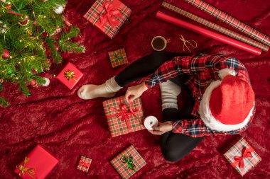 Kırmızı Noel Baba şapkalı bir kadının, Noel ağacının yanındaki kabarık ekose kumaşta oturan aile için Noel hediyesi kutularını paketlediği en iyi manzara. Düz yatıyordu.