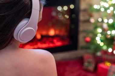 Kulaklıklı kadın kış akşamı şömine ateşi ve yılbaşı ağacının yanında oturup ısınıyor..