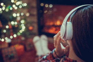 Kulaklıklı bir kadın kış akşamı şömine ateşi ve noel ağacının yanında oturup ısınıyor..