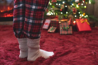 Pijama giymiş kadın ayakları ve kış çorapları Noel ağacının yanında duruyor. Hediyeler ve şömineyle birlikte. Kavram