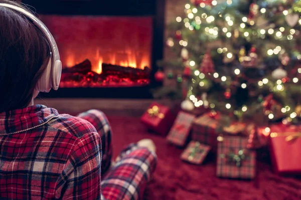 暖炉の炎とクリスマスツリーの近くの冬の夜に座って暖まるヘッドフォンで一人で悲しい女性 — ストック写真