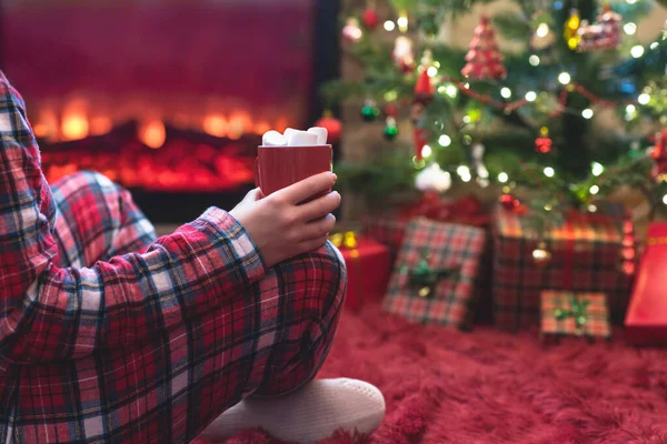 冬天的晚上 穿着睡衣 拿着热可可和棉花糖的女人坐在壁炉边和圣诞树旁取暖 — 图库照片