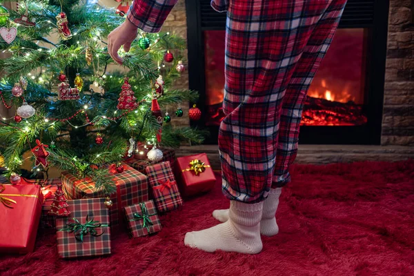 Woman in pajama and socks decorating branches of christmas tree with toy at home near fireplace.