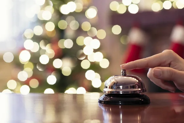 Hand of guest ringing reception bell on desk of guesthouse, hotel at christmas time. Color shining garland on christmas tree on background. Travel concept.