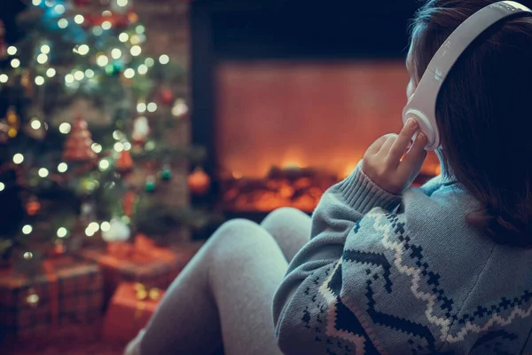 冬天的晚上 戴着耳机的女人坐在壁炉边和圣诞树旁取暖 — 图库照片