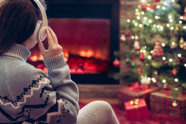 暖炉の炎とクリスマスツリーの近くの冬の夜に座って暖まるヘッドフォンの女性 — ストック写真