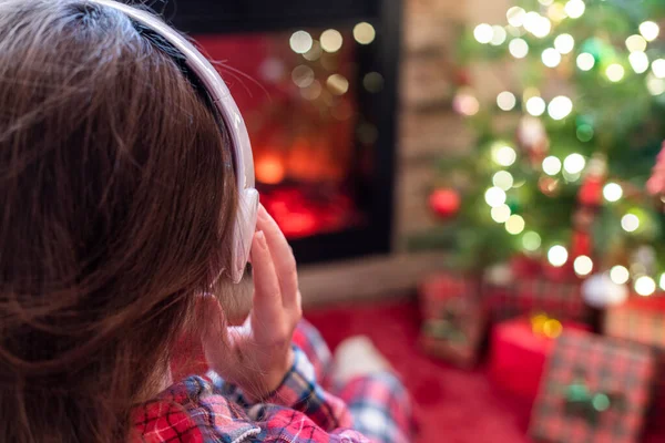 冬天的晚上 穿着睡衣 头戴耳机的女人坐在壁炉边和圣诞树旁取暖 — 图库照片